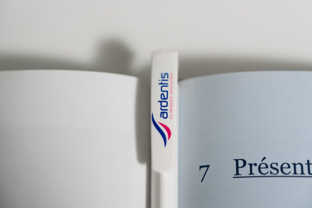 Close-up of a white Ardentis branded pen resting on an open book, highlighting office supplies and branding.