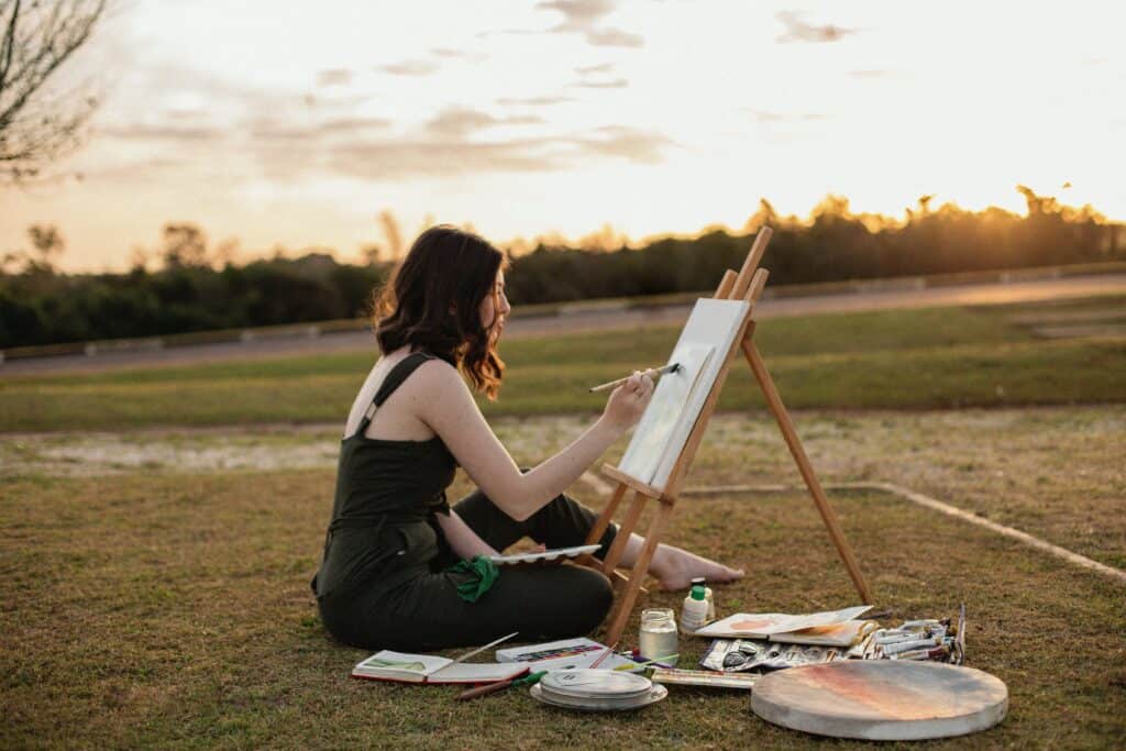 A woman sitting on the grass, painting on a canvas with an easel during sunset, surrounded by art supplies in an open field.