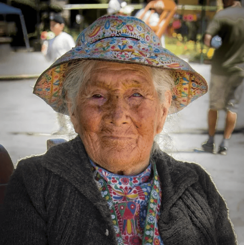 Elderly woman with a colorful hat and patterned clothing enjoying an outdoor event, smiling at the camera.