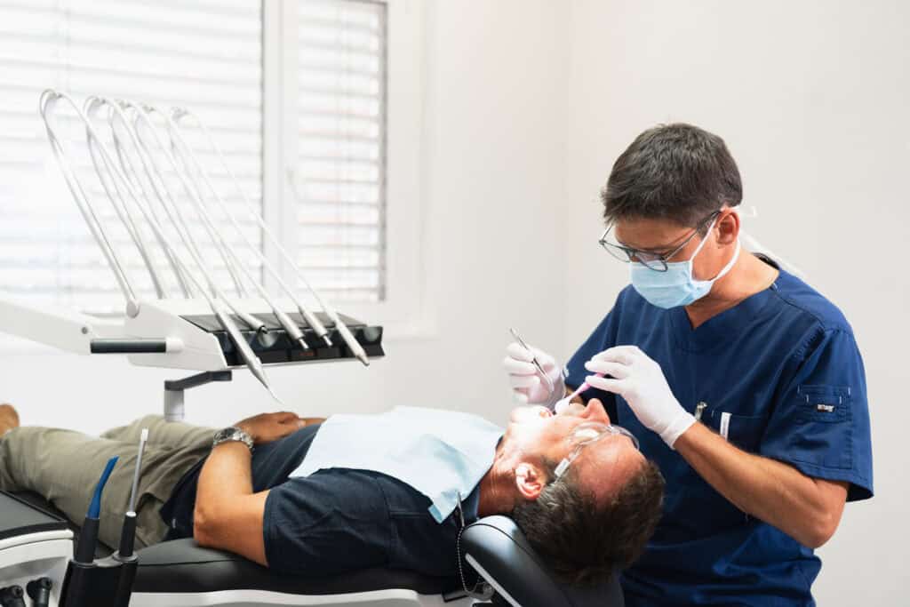 A dentist in protective gear performing a dental procedure on a patient lying in the dental chair.