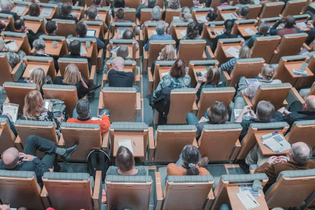 Audience attending an Ardentis conference in a large auditorium, seated in rows with notebooks and devices, engaging with the presentation.
