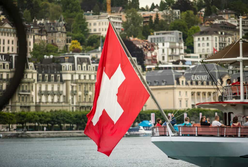 Swiss flag flying on a boat docked at a city waterfront with historic buildings and lush greenery in the background.