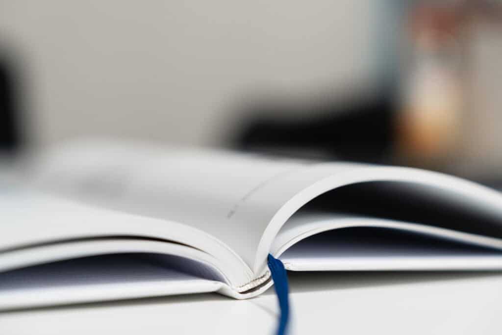 A open book with white pages and a blue ribbon bookmark, set against a blurred background, representing reading, learning, or documentation.