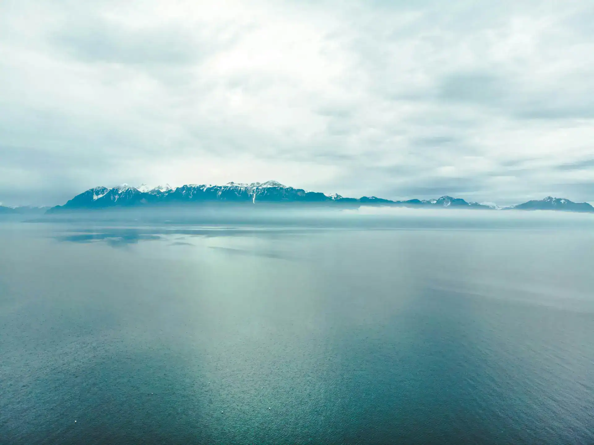 Tranquil lake with distant snow-capped mountains under a cloudy sky, showcasing natural beauty and peaceful scenery.