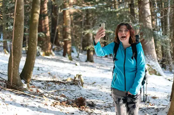 Femme souriante en randonnée dans une forêt enneigée, utilisant son smartphone pour prendre une photo, portant un manteau bleu et un sac à dos.