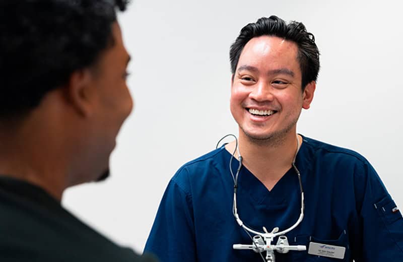 Dentiste souriant en consultation avec un patient, portant une blouse bleue et un stéthoscope, dans un cabinet dentaire moderne Ardentis.