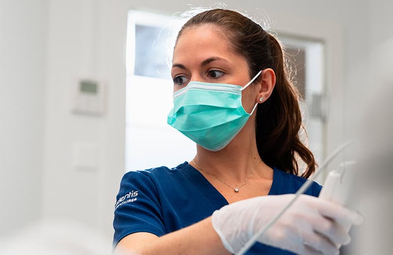 Praticienne dentaire portant un masque et des gants, utilisant un instrument dentaire dans un environnement clinique propre et professionnel.