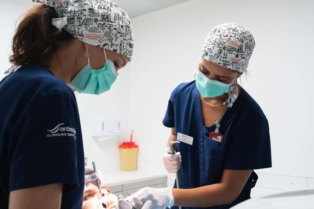 Two dental professionals in scrubs and masks providing dental treatment to a patient in a clean, modern clinic environment.